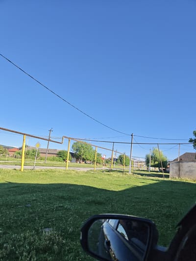 The image shows a sunny day in a rural village, likely Mayrtup, Russia. The scene is viewed from the perspective of someone inside a car, as indicated by the side-view mirror in the foreground. The sky is a clear, bright blue, suggesting it is daytime.
The landscape is dominated by green grass and a few trees. Yellow poles with horizontal pipes, possibly part of a fence or utility structure, run across the middle ground. There are also power lines and utility poles visible. In the background, there are houses with tiled roofs and some farm animals, possibly cows, grazing. A road is visible with a yield sign. The overall impression is one of a peaceful, rural setting on a pleasant day. There is no visible text in the image.