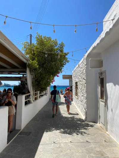 The image depicts a sunlit, narrow, stone-paved alleyway leading towards a coastline in a Greek town, identified as Φάρος. The alley is flanked by whitewashed buildings. On the left, an outdoor dining area under a wooden canopy, with visible patrons and a low white wall, abuts a large green tree with pink floral accents. On the right, a textured white stone building features a recessed opening and a closed, light-colored wooden door. Overhead, multiple strings of bare bulb lights and utility lines are visible against a clear blue sky. In the foreground left, a female subject in sunglasses and light-colored clothing is observed interacting with a mobile phone. In the mid-distance, two subjects, a male and a female, are walking away from the viewer towards the ocean. The male wears a dark shirt, patterned shorts, and carries a light blue tote bag. The female wears a colorful patterned dress and a red hat. The background reveals a turquoise sea, a sandy beach with indistinct figures, and several large white yachts. Shadows are cast upon the pavement from the structures and tree.