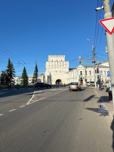 The image captures a view of a street in the city of Feklino, Russia, on a clear, sunny day. In the background, a large, white, crenellated structure, likely a historical gate or tower, dominates the scene. In front of this structure, a group of people are visible near an arched entryway. To the right of the tower, a white building with multiple windows is situated.
The foreground of the image shows a paved road with white lane markings. Several cars are present on the road; a dark SUV is parked on the left side, and a silver sedan is driving away from the viewer in the center-right. A crosswalk is marked on the road.
Along the right side of the road, there is a sidewalk with several pedestrians. A tall, gray utility pole with various wires, a security camera, and traffic signals is prominently featured. A red and white triangular road sign, indicating a yield or give way, is attached to this pole. Other road signs, including a blue directional sign with an arrow and a white sign with the word "СТОП" (STOP in Russian), are also visible.
To the left of the road, there are several tall evergreen trees. Traffic lights are also visible, with one showing red. The sky is a vibrant, clear blue. The overall impression is of an urban environment with historical architecture and active street life.