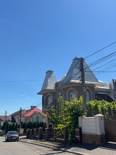 The image depicts a residential street in the city of Chahor, Ukraine, under a clear blue sky. A dark-colored car is parked on the left side of the asphalt road, facing away from the viewer. Several houses are visible along the street, with architectural styles varying. The house in the foreground on the right features a prominent, dark gray facade with large arched windows and a steeply pitched, silver-colored roof. A utility pole with numerous electrical wires stands in front of this house. To the left, other houses are visible with different colored roofs, including one with a red tile roof. Fencing and lush green trees and bushes line the properties. The overall scene suggests a quiet suburban or residential neighborhood.