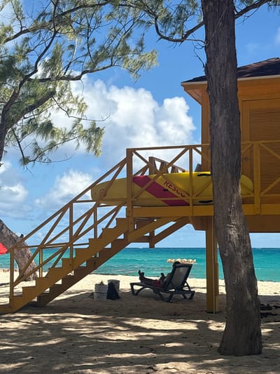 This image captures a beach scene in Wellhouse, Barbados, under a clear sky with scattered white cumulus clouds. In the midground, a two-story yellow lifeguard station stands on the sandy beach. The station features a brown shingled roof and an enclosed upper level with louvered panels. A yellow wooden staircase with railings ascends to a landing on the first level. A yellow rescue paddleboard, marked with "RESCUE" in red capital letters and a diagonal red stripe, is stored horizontally on this landing.
To the left of the station, partially obscured by a tree, a red umbrella is visible on the sand. Directly beneath the staircase, a single person lies reclined on a dark beach lounge chair, facing the ocean. The individual wears red shorts and is actively engaging with a mobile device held in their hands, with their feet elevated on the chair. To the left of the chair, two white buckets and a dark bag rest on the sand.
The foreground is dominated by light-colored sand, marked with dappled shadows from several trees, likely casuarinas, whose trunks and needle-like branches are visible on the left and right sides of the frame. Beyond the beach and the lifeguard station, the turquoise-blue ocean stretches to the horizon, with small waves visible in the distance. A small, flat, rectangular object floats on the water's surface further out.