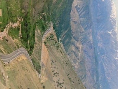 An aerial view of a winding road in a mountainous region of Uzbekistan, captured from a high vantage point. The road winds through a steep, heavily forested hillside, with sparse vegetation. The road is partially obscured by a shadow, indicating the time of day as late afternoon or early evening. Several vehicles are visible travelling on the road. The image is taken from a location at latitude 40.861, longitude 71.46 in Kalgan-Dar'ya. The hillside is covered in trees, some of which are deciduous. The road curves gently to the right, disappearing around a bend. The image is taken from a distance, with the road and hillside appearing small in the frame. The sky is obscured by hazy clouds. The mountains in the background are distant and hazy. A thin layer of snow covers the distant mountain peaks. The image does not show any people.