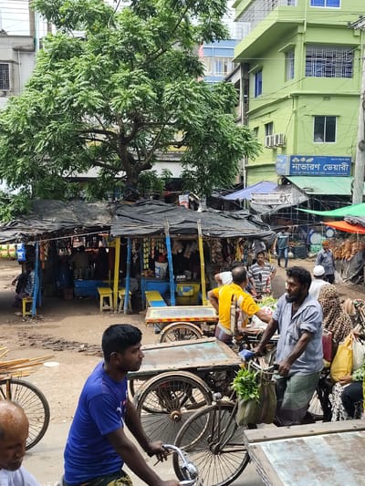 The image depicts a bustling street scene in Benapole, Bangladesh, likely a marketplace given the presence of numerous stalls and vendors. In the foreground, a man in a blue t-shirt is pushing a bicycle laden with goods. To his right, another man in a striped shirt is also operating a bicycle cart, which has bags of green leaves hanging from the handlebars. Several other individuals are visible in the mid-ground and background, engaged in various activities such as tending to stalls or walking through the crowded area. The stalls are covered with tarps, displaying a variety of items, including packaged goods and fresh produce. The environment is characterized by a mixture of commercial activity and everyday life, with buildings and trees forming the backdrop. The ground appears to be unpaved, suggesting a typical street in a densely populated urban area.