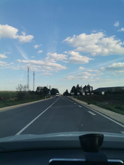 The media file presents a view from inside a vehicle, looking forward down an asphalt road on a clear day with scattered white cumulus clouds against a bright blue sky. The time of day appears to be late afternoon or early evening due to the soft lighting.
The road features a solid white line dividing the lanes and broken white lines along the right shoulder. To the left, the landscape consists of green fields, some patches of dark, cultivated earth, and two tall metal electricity pylons supporting power lines that traverse the scene. Further left, trees and what might be agricultural or industrial structures are visible, with a small red tractor partially obscured in the far distance.
On the right side of the road, there is a gravel shoulder, followed by a line of evergreen trees. Beyond the trees, several white container-like structures are present, backed by a dark, elevated landform, possibly a hill or a spoil heap. An oncoming white van with its headlights on is visible in the distance.
The immediate foreground shows the dashboard of the car, with a dark, rounded object, likely a dashcam or sensor, mounted on it. A small, white road sign on the right, partially hidden by trees, bears the Cyrillic text "C. ЦАРКВА," indicating the location as Tsarkva, Bulgaria. The scene depicts a drive through a rural or semi-rural area. No individuals are explicitly visible, though the white van suggests other road users.