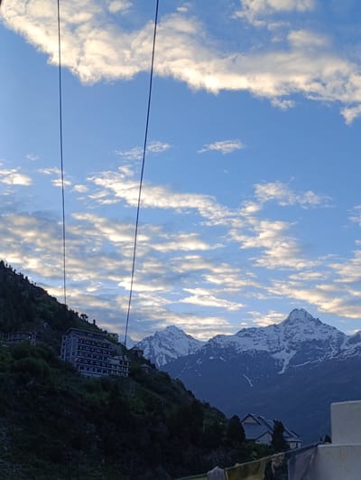 This is a photograph taken in Kardang, India, featuring a dramatic landscape under a cloudy sky. In the foreground, two dark, thin wires or cables stretch diagonally across the frame from the top left to the bottom right, obscuring parts of the scene behind them.

Dominating the mid-ground is a steep, green mountainside. Perched precariously on this slope is a large, multi-story hotel building. The hotel is light gray and appears to have many windows, with some lights illuminated, suggesting it is occupied. The word "HOTEL" is visible in capital letters on the facade. The mountainside is covered with dense vegetation, including trees, and a few smaller buildings or structures are scattered around the hotel.

In the background, a range of majestic snow-capped mountains rises. These peaks are sharp and rugged, with visible snow cover and shadowed crevices. The sky above is a vibrant blue, dotted with numerous white and gray clouds, some of which are illuminated by what appears to be early morning or late afternoon sunlight, creating a textured and dynamic ceiling to the scene. The overall impression is one of a remote, high-altitude location with dramatic natural beauty and human infrastructure built into the challenging terrain.