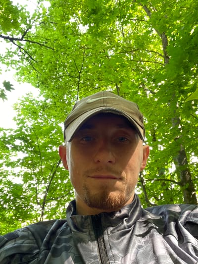This is a portrait-oriented, low-angle, close-up shot of a man looking up at the camera, with lush green trees and foliage filling the background. The man is wearing a baseball cap and a dark, patterned jacket with a zipper. He has short, light-colored hair and stubble. His face is partially obscured by the brim of his cap and the angle of the shot. The leaves of the trees are vibrant green, suggesting it is daytime and likely a warm season. Sunlight filters through the leaves, creating a dappled effect. The overall impression is one of being outdoors, possibly in a forest or park, with the camera positioned beneath the man, looking upwards. There is no visible text in the image. The scene evokes a sense of casualness and connection with nature.