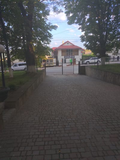 The image depicts a paved walkway leading towards a gated entrance of a building with a red-tiled roof. Two white cars are parked in front of the building, which appears to be a store with signage in Cyrillic. Tall trees with dense foliage frame the scene, and the sky above is partly cloudy. The walkway is constructed of interlocking brick pavers, and a low stone wall runs along the right side of the walkway, separating it from a grassy area. The overall scene suggests an entrance to a commercial establishment in Zalishchyky, Ukraine, possibly a grocery store, indicated by the Cyrillic text on the building's facade.