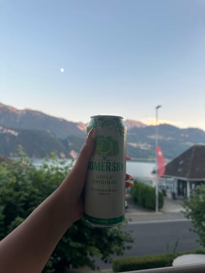 A hand holds a can of Somersby Apple Original cider. The can is predominantly white with green decorative patterns and text. Visible text on the can includes "SOMERSBY", "APPLE ORIGINAL", "REFRESHINGLY CRISP", and "ALC. 4.5% VOL". The background depicts a scenic mountainous landscape with a body of water and a building. The sun appears to be setting or rising, casting a warm glow on the mountains. The sky is a clear blue with a small, faint moon visible. Lush green trees are present in the foreground and mid-ground, suggesting a temperate climate. The overall impression is one of a relaxing moment enjoyed outdoors in a beautiful natural setting, likely in the late afternoon or early evening. The image is oriented vertically.