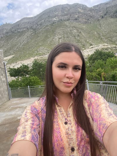 A young woman with long, dark hair and dramatic false eyelashes takes a selfie outdoors. She is wearing a short-sleeved button-up shirt with a colorful, abstract pattern in shades of pink, orange, and purple. Behind her, a large, barren, rocky mountain slopes upward, sparsely covered with green vegetation and dotted with a few trees. A metal railing runs horizontally across the middle ground. To the left, a portion of a textured, grey brick building is visible, and the ground appears to be a concrete surface with white lines painted on it. The sky above is cloudy. The location is identified as Chalda, Russia, though this specific scene does not contain any discernible urban elements that would confirm it is within a city.