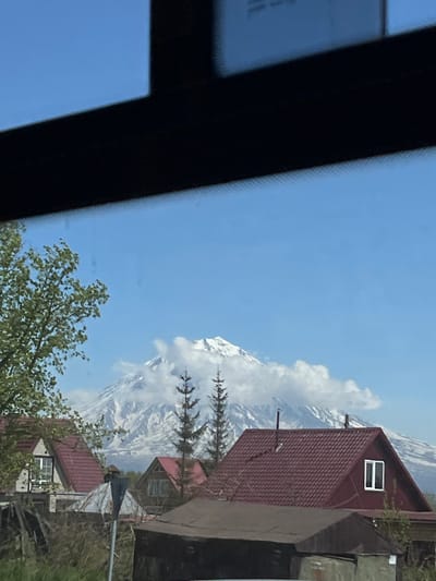 A frontal, eye-level shot depicts a residential area in front of a snow-capped mountain under a clear blue sky. In the foreground, a black, trapezoidal object, possibly a car visor or frame, occupies the top portion of the image. Below this, to the left, is the upper canopy of a green deciduous tree with a textured bark.
The middle ground features several houses with red, shingled roofs, some with visible windows and chimneys. Among these structures, a gray, pole-like object with a triangular top is prominently positioned. Two evergreen trees are situated between the houses and the mountain.
The background is dominated by a majestic, snow-covered mountain with billowing white clouds partially obscuring its peak. The base of the mountain extends into the distance, hinting at a vast landscape. The scene appears to be a daytime setting. The geographical location is specified as Krasny, Russia, suggesting a rural or semi-rural environment.