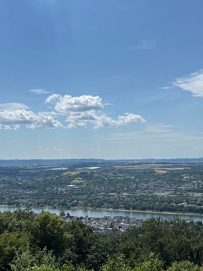 This is a vertical, high-angle, medium shot of a landscape view. The scene features a body of water, likely a river, flowing through a valley. On the left bank of the river, a densely populated town or city stretches along the water's edge, characterized by numerous buildings and interspersed with lush green trees. The right bank of the river also shows signs of habitation, with more buildings visible amidst the greenery. In the foreground on the right, a dense collection of trees with dark green foliage occupies a significant portion of the frame, creating a natural border.

The background reveals a hazy, expansive landscape with rolling hills or mountains fading into the distance. The sky above is a clear, bright blue, dotted with scattered white clouds, some of which are wispy and elongated, while others are more puffy and voluminous. A distant hot air balloon is visible in the sky above the town.

The lighting suggests it is daytime, likely late morning or early afternoon, given the brightness and clear sky. The weather appears to be fair and sunny. The scene evokes a sense of tranquility and offers a panoramic view of a European settlement nestled within a natural environment. No people or text are discernibly visible. The image captures a scenic vista of Königswinter, Germany, with its characteristic riverside setting.