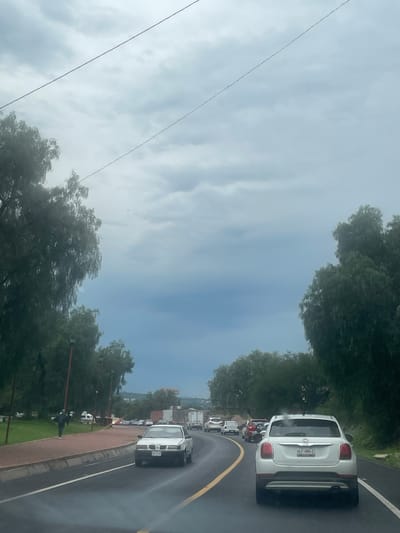 The image depicts a cloudy, overcast day on a highway near Tepeji de Ocampo, Mexico. The perspective is from within a vehicle, looking forward. Several cars are on the road, including a white Fiat 500 in the foreground, a white sedan to the left, and various other vehicles in traffic ahead. To the left of the highway, there is a sidewalk with a person walking, lined by trees and lampposts. The right side of the road is bordered by trees and a slight embankment. In the distance, the landscape appears hilly and somewhat arid, with some buildings visible. Overhead, power lines are strung across the sky, and faint red and pinkish lights are visible against the cloudy sky, possibly lens flares or atmospheric phenomena. The overall impression is that of a typical cloudy day with traffic on a highway.