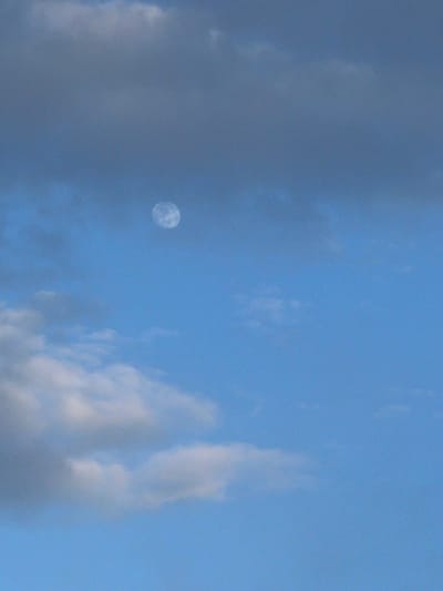 The image captures a daytime sky, predominantly blue, with varying cloud formations. A pale, circular moon is visible towards the left-center of the frame, showing faint surface details. The sky transitions from a brighter, lighter blue on the right side to a slightly deeper, more muted blue on the left, influenced by the presence of clouds. The clouds themselves are diverse, ranging from wispy, bright white patches to denser, darker grey masses, particularly on the left side. The scene depicts the natural phenomenon of the moon being visible during daylight hours. There are no people, man-made objects, or discernible text present. The weather appears partly cloudy. The context provided indicates the location is Hudson, United States.
