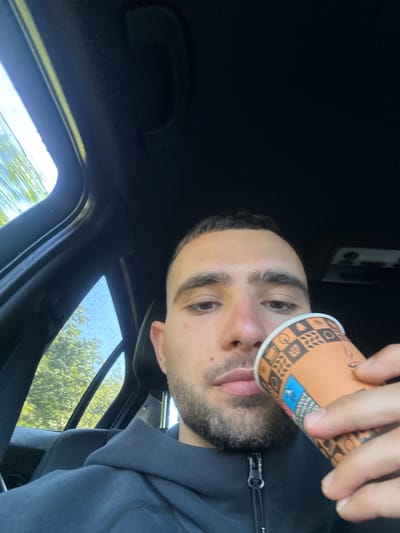 The image is a close-up, angled selfie of a young man taken inside a vehicle. The man, likely in his 20s or 30s, has short dark hair, distinct eyebrows, and a trimmed beard or stubble. He is wearing a dark hooded jacket with a prominent silver zipper. His face is angled towards the camera, with a neutral to slightly contemplative expression.

He holds an orange and brown paper cup with his right hand, positioning it near his mouth as if about to drink or having just taken a sip. The cup features various decorative patterns including coffee beans, mugs, and geometric shapes.

The setting is the interior of a car or similar vehicle, with the dark headliner filling much of the upper left portion of the frame. Through the windows on the right and bottom, bright green foliage from trees and glimpses of a bright sky are visible, indicating an outdoor, daytime scene. Small water droplets are noticeable on the window panes, suggesting recent rain or condensation. There is a grab handle or similar car interior feature visible on the left side of the frame. No legible text is visible on the cup or in the background. The location context is Хаджийско, Bulgaria.