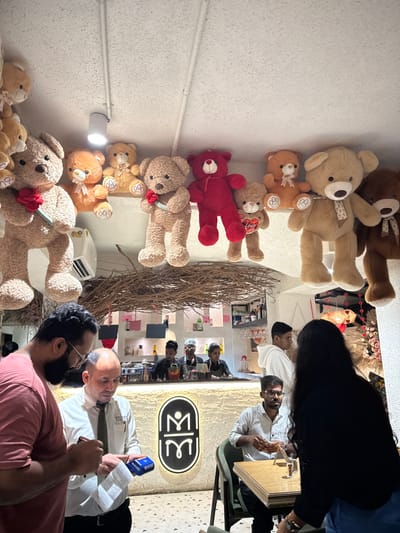 The image depicts the interior of a commercial establishment, likely a cafe or restaurant, located in Kalyan-Dombivli, India. The scene features multiple individuals and a distinctive decor dominated by stuffed teddy bears.
In the foreground, two men are engaged in a transaction. On the left, a man with a beard and glasses, wearing a salmon-colored t-shirt, holds a smartphone or similar device in his hands. Facing him, a man with a bald head, wearing a white collared shirt and a dark tie, holds a white paper receipt and a blue payment terminal. Behind them, a prominent illuminated logo, consisting of stylized interlocking 'M' shapes, is mounted on a light-colored, textured wall.
To the right, a man with glasses and a short beard, dressed in a light-colored checkered shirt, is seated at a light-colored wooden table with a beverage glass, looking towards the left. Partially obscuring him, a person with long dark hair, seen from behind, wears a black top and blue jeans.
In the mid-ground, a brightly lit bar counter is visible, behind which four individuals are present. Three staff members in dark uniforms are engaged in preparing drinks, with bottles, glasses, and a coffee machine visible on the counter. Another individual in a white hoodie stands behind the counter, looking right.
The upper portion of the scene is heavily decorated with numerous stuffed teddy bears of various sizes and colors (light brown, dark brown, red, orange), suspended from the ceiling or placed on elevated ledges. Several bears hold small red objects, such as roses or hearts labeled "LOVE." Below these decorations, a lattice-like structure made of intertwined branches or twigs spans across the ceiling. A white wall-mounted air conditioning unit is visible on the upper left, beneath some teddy bears. The floor is a light-colored terrazzo.