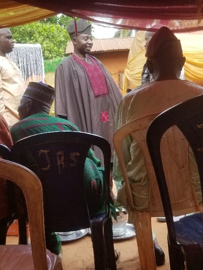 The image depicts a social gathering under a brown translucent canopy in Ochadam, Nigeria. In the foreground, two men are seated with their backs to the viewer. The man on the left occupies a dark plastic chair with "JRS" inscribed on its back, wearing a green and black patterned tunic and a dark cap. The man on the right is seated on a light beige plastic chair, wearing a light green tunic. Between them, two small metal buckets rest on a metallic tray on the earthy ground.
In the mid-ground, a standing man, positioned slightly left of center, is smiling and looking towards the viewer's right. He wears a long, dark olive-brown traditional tunic with a burgundy embroidered chest panel and a matching embroidered design near the hem, complemented by a dark green cap with a distinctive upward curve. To his left, another man is partially visible, wearing a light tan traditional outfit.
The background features green foliage, a structure with a corrugated metal roof, and yellow fabric draping, suggesting decorative elements. The scene is illuminated by natural daylight.