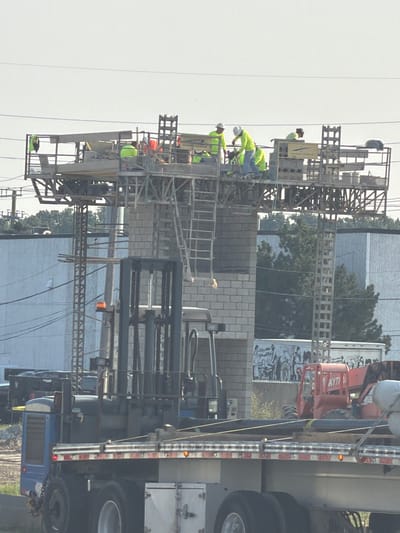 In Shelby Charter Township, Michigan, United States of America, a construction scene unfolds with workers on a platform above a brick structure. Five individuals, all wearing high-visibility safety vests, are actively engaged in their tasks on the elevated platform. Two workers are dressed in yellow vests, while three are in green vests. One worker in a yellow vest is leaning over, appearing to be working with tools, while another in a green vest is also bent over, potentially operating machinery or handling materials. The platform itself is a complex structure of scaffolding and beams, with a ladder providing access. Below the platform, a large forklift is positioned prominently, its mast raised, indicating it might be in use or ready for operation. In the foreground, a flatbed truck carries several long, dark pipes secured for transport. Behind the forklift and truck, a red piece of heavy machinery, possibly a boom lift, is visible, and further back, a white trailer with an image of people is parked. The overall environment suggests an active construction site, with buildings and trees forming the backdrop under a clear sky.