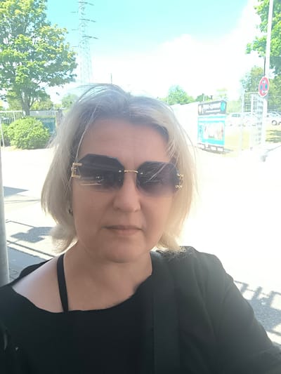 An outdoor selfie shows a woman with shoulder-length blonde hair and sunglasses standing in front of a blurred background. She is wearing a black top. The background includes trees, a power pylon, fences, a sign with text that appears to be in German, and a street. The lighting suggests it is daytime with bright sunlight. There is no indication of specific interactions or actions beyond the selfie being taken. The location is identified as Eschborn, Germany.