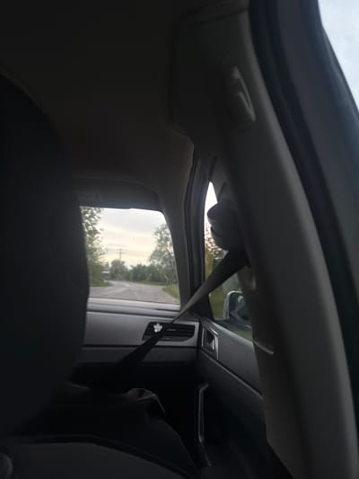 The image shows the interior of a car, specifically the driver's side, looking slightly towards the back. The view is partially obscured by the back of the driver's seat. The open window shows a glimpse of the outside, including trees and a road. A seatbelt is visible. The car's dashboard and interior trim are also visible. It is difficult to place the location in Krainitsi, Bulgaria without more information.