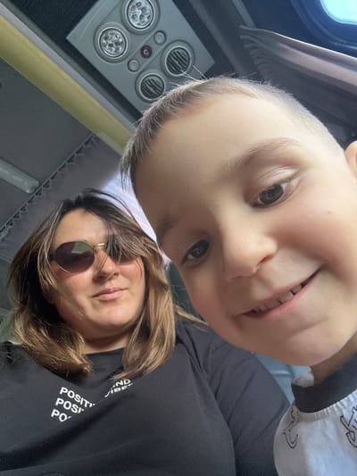 This is a close-up, low-angle selfie taken inside a vehicle, possibly a bus, with the roof lights and overhead compartments visible in the upper portion of the frame. The photo features two individuals: a woman in the foreground on the left and a young boy on the right, whose face is prominently displayed. The woman is wearing sunglasses and a black t-shirt with white text that reads "POSITIVITY AND POSITIVE VIBES". Her hair is brown and appears to be windswept. The boy, positioned closer to the camera and slightly out of focus, has light brown hair and is smiling, showing his teeth. The interior of the vehicle is dimly lit, with a yellow handle bar running horizontally across the frame. The overall impression is a candid moment captured during travel, though the specific location in Ivanovka, Russia cannot be determined from the visual cues alone.