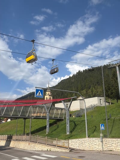 The image captures a scene in Pozza di Fassa, Italy, with a cable car system overhead and a pedestrian crossing on the road below. Two cable cars, one predominantly yellow and the other gray, are suspended from wires against a blue sky dotted with white clouds. In the middle ground, a bridge with a red net canopy spans across a grassy slope, leading to a building with white walls. A church tower with a yellow spire and clock is visible behind the bridge. A blue pedestrian crossing sign is prominently placed near the road. The foreground shows a paved road with white markings for a pedestrian crossing, bordered by a stone wall and a wooden fence. The background features a densely forested mountainside. The overall atmosphere is serene and picturesque, typical of a mountain resort town.