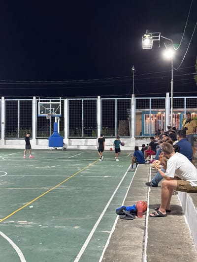 A nighttime scene shows a basketball court with two hoops and various people. Along the side are spectators watching the action. Several individuals are on the court, either dribbling basketballs or standing around. Gear and personal items rest along the sideline. A bright light illuminates the scene, creating a strong contrast between the lit court and the dark surroundings. The fence around the court is partially visible.