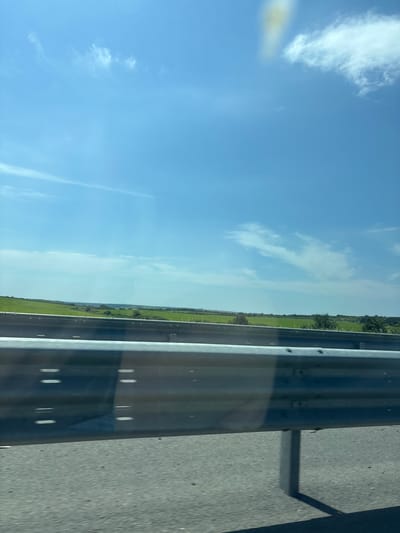 A highway guardrail is in the foreground, with a gray asphalt road surface visible below it. Beyond the guardrail, a vast expanse of green fields stretches to the horizon. The sky above is a clear, bright blue, dotted with scattered white clouds. A slight yellow tint is visible in the upper right portion of the sky, possibly from a reflection or lens flare. The scene appears to be a landscape captured from a moving vehicle on a highway, likely in or near Malko Tarnovo, Bulgaria, given the characteristic rolling green hills often found in the Strandzha region. No people or specific buildings are identifiable.