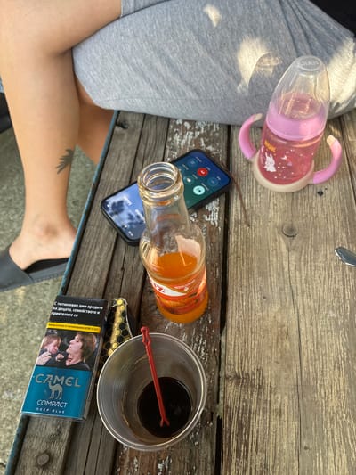 A high-angle outdoor shot captured in Radievo, Bulgaria, shows a weathered wooden plank table laden with various items, with the lower body of a person seated behind it. The individual wears grey shorts or sweatpants, with one leg crossed over the other. A dark tattoo, resembling a bird or feather, is visible on their left lower leg, which also wears a dark grey slip-on sandal.

On the table, from left to right, are:
*   A blue "CAMEL COMPACT DEEP BLUE" cigarette pack, featuring a graphic health warning in Bulgarian depicting a crying child and an adult exhaling smoke.
*   Next to the cigarette pack, a small object with a textured, golden-yellow and black honeycomb-like pattern, possibly a cigarette case or lighter.
*   A clear plastic cup containing a dark brown liquid and a red plastic stir stick.
*   A black smartphone, face-up, displaying a call screen with Cyrillic text and a "00:00 / 00:00" timestamp. The screen reflects ambient light.
*   An amber glass bottle, partially filled with an orange liquid, resting beside the smartphone. Its label is partly visible, showing "UNITEME" and "200ml" text.
*   A pink NUK baby bottle with handles, containing a pink liquid and decorated with white rabbit decals, positioned on the right side of the table.
*   The tip of a silver spoon is visible on the far right edge of the table.

The wooden table surface exhibits prominent grain and wear, with chipped paint in areas. Sunlight casts shadows, notably on the person's clothing.