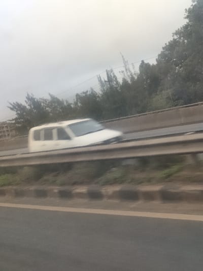A forensic examination of the image reveals an outdoor scene depicting a multi-lane road environment under an overcast sky, consistent with the Kahawa Sukari ward, Kenya. The perspective indicates the photograph was captured from within a moving vehicle, looking across to a parallel road.
In the immediate foreground, a dark gray asphalt road surface is visible, separated by a light-colored line from a concrete curb containing dark rectangular blocks. Beyond this curb, a two-bar metal guardrail runs horizontally across the mid-section of the image. Directly behind the guardrail, a light-colored multi-purpose vehicle (MPV) or sport utility vehicle (SUV) is in motion from left to right. The vehicle, primarily white or light gray, is heavily motion-blurred, suggesting a significant speed relative to the camera. A roof rack is discernible on the vehicle's roof.
Behind the vehicle and its lane, a dense line of dark green trees and shrubs forms a visual barrier against the horizon. Some taller, thinner tree silhouettes are visible further back. Faint, thin lines, consistent with utility wires, traverse the upper background horizontally above the treeline. The sky is uniformly light gray, indicating heavy cloud cover. No individuals are visible in the frame. The overall impression is a highway or major road on a potentially hazy or overcast day.