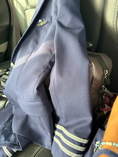 The image shows a blue suit jacket with three stripes on the sleeve and a winged pin on the lapel, lying on what appears to be a car seat. A bag with a zipper is visible next to the jacket. The location is not discernible.