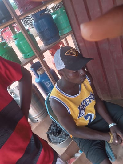 A medium shot, taken at eye level and rotated clockwise, depicts a man seated among a rack of pressurized gas tanks in Eka, Nigeria. He wears a yellow Los Angeles Lakers jersey and a black cap with a mesh back. He has two wristbands, one white and one yellow. A red and black striped shirt is visible in the lower right corner, and the top of a silver keg or container is to its left. More gas tanks are visible in the wooden rack.