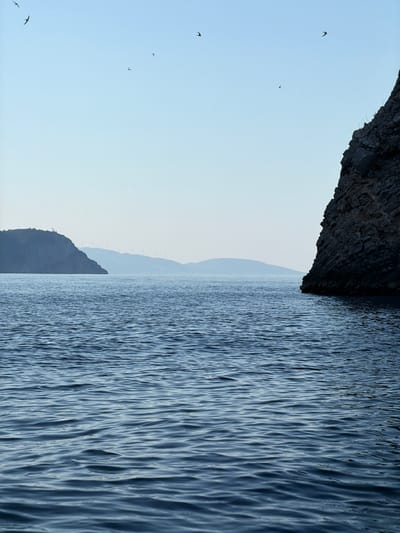 A wide-angle, eye-level shot captures a serene seascape with a pale blue sky and calm, rippling water in the foreground. On the left side of the frame, a silhouetted, dark, mountainous landmass is visible in the distance, dotted with wind turbines. A sheer, dark rock face dominates the right side of the frame, rising from the water. Several small, dark birds are silhouetted against the sky. The scene is devoid of human presence. The image is a landscape photograph taken from the water looking towards the coast of Prijevorac, Montenegro.