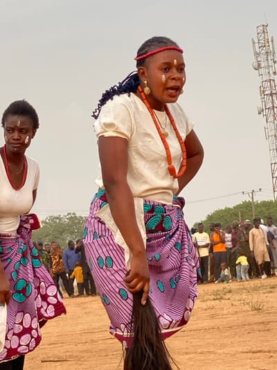 This image captures a vibrant cultural event in Agunu, Nigeria, showcasing two women in traditional attire performing outdoors before an audience.
The primary subject, closer to the viewer, is a young woman wearing a light-colored, short-sleeved top and a colorful patterned wrapper (skirt) featuring lavender, teal, and maroon/brown hues. She is adorned with a prominent red beaded necklace and a matching red headband or braided accessory. Small, circular gold earrings complement her ensemble. Her face bears distinct white markings on her forehead, cheeks, and chin. She has a dynamic expression, with her mouth slightly open, suggesting she is singing, speaking, or engaged in a lively performance. Her dark hair is styled, possibly in braids or twists. Her right hand holds a long, dark object, which appears to be a traditional whisk or horse tail, extending out of the frame.
Behind her and slightly to her left, a second woman is partially visible, dressed in similar attire—a light top, a patterned wrapper, white face markings, and a red beaded necklace. She looks towards the camera with a more composed expression.
In the background, a crowd of spectators, consisting of adults and some children, stands in rows, observing the performance. They are dressed in a mix of casual and traditional clothing. The setting is an outdoor area with a light brown, dusty ground. Sparse green foliage or trees are visible in the far background. A metal lattice tower, resembling a communication tower, stands prominently against the light, possibly overcast or hazy, sky in the upper left section of the image. The scene suggests a daytime event, filled with cultural significance and community participation. No visible text is present.