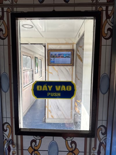 The image shows a close-up of a glass door with a sign that says "ĐẨY VÀO PUSH" in yellow lettering on a blue background with a yellow border. The sign is located on the right side of the door. The door appears to be a part of a building or vehicle, possibly a train car, given the view through the glass. The interior of the building/vehicle is visible, showing windows and decorative elements on the walls. The exterior of the door is decorated with a stained-glass-like pattern. There is a metallic handle visible on the bottom left of the image. The overall impression is of an entrance with a directional sign. There are no people visible in the image. There is no information in the image to identify the location as Bình Long, Vietnam.