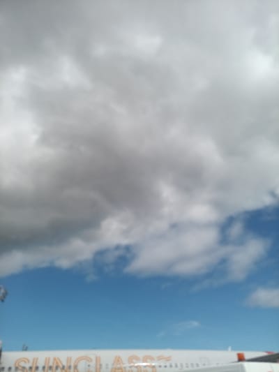 The image displays an outdoor scene in Ojos de Garza, Spain, centered on the upper portion of a white passenger aircraft under a mixed cloudy and blue sky. The aircraft's fuselage, visible in the lower frame, features orange lettering that partially spells "SUNGLASS" and an orange stripe near its rear. Above the aircraft, the sky transitions from a predominantly grey, overcast cloud layer in the upper half to a clear blue sky with some scattered white clouds in the lower middle and right. No persons or explicit actions are discernible. The image is generally blurry.