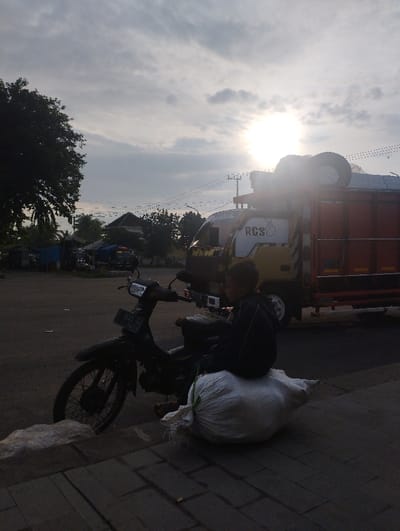 In a subdued light, likely early morning or late afternoon, a young boy is captured sitting on a large white sack next to a parked black moped-style motorcycle. He is wearing a dark jacket and is positioned facing away from the camera, giving a sense of waiting or rest. The motorcycle's license plate, "P 5025 NM" with "07 26" below it, is visible.
In the mid-ground, a yellow and orange truck is prominent, with tires stacked on its roof or top section. The text "RCS" along with what appears to be a heart symbol is clearly visible on the side of the truck's cab. The sun, a bright glare, is low in the sky, positioned in the upper right, casting soft light and suggesting the time of day. The sky is partly cloudy, with diffused light.
To the left, dense green trees frame the scene, and power lines are visible across the distant horizon. The ground beneath the boy and motorcycle is paved with asphalt, transitioning to concrete pavers in the foreground. The overall scene depicts a quiet, possibly mundane moment by the roadside in Kecamatan Jangkar, Indonesia.
