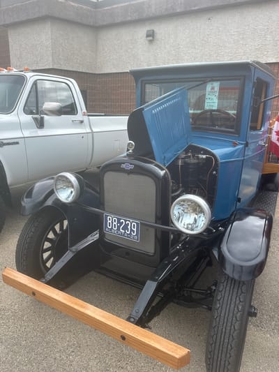 A blue vintage Chevrolet truck with its hood open is parked on asphalt in front of a light-colored, textured building. To the left of the Chevrolet, a white pickup truck is partially visible. The Chevrolet has a black grille with a Chevrolet emblem, two large round headlights, and a license plate that reads "88-239 ALBERTA-28". A wooden beam is mounted at the front of the Chevrolet's frame. Inside the cab, a windshield wiper is visible, and a paper is attached to the windshield, indicating "Canada Day Car Show" and "Vintage" in the award category. A Canadian flag is attached to the right side of the truck.