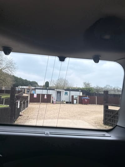 The image, captured from the interior of a vehicle looking out through its rear window, depicts an outdoor compound or facility under an overcast sky in Denford, United Kingdom. The vehicle's dark grey headliner and rear window trim are visible at the top and bottom of the frame, with visible defroster lines across the window.
The exterior scene features a gravel or dirt ground covering a large open area. Several single-story modular buildings with light grey or off-white walls and dark roofs are present. A prominent modular building in the center-right has a dark grey door, a small window, and potted plants near its entrance. Surrounding the open area and structures is extensive dark brown wooden fencing, including sections with decorative tops and a distinct wooden gate on the far right. Attached to a fence section to the left of the central modular building are two light-colored signs, one featuring red text. To the right of the building, a reflective sign or mirror is visible near a smaller dark brown bin. Further back on the right, a large dark red waste bin with a blue lid stands behind the fencing. The background shows deciduous trees without leaves and some evergreen foliage, suggesting a colder season. No individuals or active interactions are present in the scene.