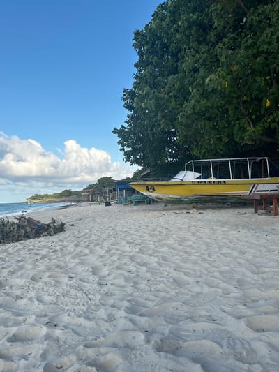 The image shows a tropical beach scene. In the foreground, white sand is textured with footprints and scattered debris. A yellow boat with a white canopy sits on the sand, partially covered by a large tree with dense green foliage. In the background, the turquoise ocean meets a coastline dotted with vegetation and several small structures, including thatched-roof huts and a covered seating area. The sky is bright blue with scattered white clouds. The scene appears to be a public beach with recreational facilities. The location is not in A R A, Indonesia, but rather a tropical coastal area.