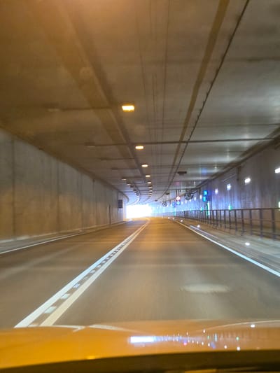 The image shows a view from the driver's perspective inside a modern tunnel. The tunnel is illuminated by overhead lights and lights embedded in the walls. The road surface is asphalt with clear white lane markings, including a dashed white line and a solid white line. The tunnel walls are made of concrete. There are metal railings along the right side of the tunnel, with signs and lights visible. The tunnel appears to be curving to the right, and the bright light at the end suggests an exit. There are no people or vehicles clearly visible in the scene. Based on the context provided in the prompt, this scene is located in the city of Kelsterbach, Germany.