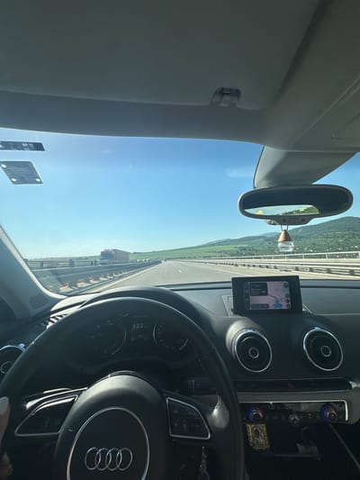 The image is a first-person perspective from inside a car, driving on a highway. The dashboard and steering wheel are visible in the foreground, indicating the car is an Audi. The car's speed is displayed as 122 km/h on the instrument cluster. A GPS navigation screen is mounted on the dashboard, showing a route. The windshield offers a view of a bright, sunny day with a clear blue sky. The highway stretches ahead, with a guardrail and other vehicles visible. In the distance, there are rolling green hills and mountains. The scene suggests travel through a rural or semi-rural area. There are no people clearly visible in the frame besides the driver's hand on the steering wheel. The location is identified as Eleshnitsa, Bulgaria.