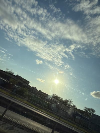 This image shows a view of the sky and a roadside from a vehicle. The sky is blue with scattered white clouds, and the sun is visible and bright. Below the sky, there is a roadside with a guardrail. Beyond the guardrail, there are buildings, trees, and vegetation. The scene appears to be a sunny day, possibly during daylight hours. The perspective suggests the photo was taken from a moving vehicle on a road, given the diagonal lines of the road and guardrail. The presence of buildings and trees suggests an urban or suburban environment. It is not possible to determine the specific location in Mölndal, Sweden from the image alone, as there are no identifying landmarks or signage.