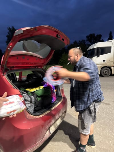 A man, smoking a cigarette, is seen from the chest down, standing next to the open trunk of a red car. He is wearing a blue and black plaid shirt and grey shorts. He is holding a colorful coiled object in his hands, which are blurred due to motion. The trunk of the car is filled with luggage, including a green duffel bag and other bags. In the background, a large white truck is parked. The scene appears to be an outdoor parking lot, possibly at dusk or night, given the dark sky. The location is implied to be Можга, Russia.