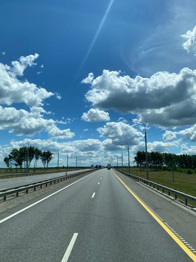 The image depicts a view from a moving vehicle on a multi-lane highway in Russia. The sky is a vibrant blue with scattered white cumulus clouds, and a faint sun glare is visible. The highway has clear lane markings and is bordered by a metal guardrail. On the left side of the road, a row of sparsely leafed trees stands behind the guardrail, with power lines and streetlights interspersed. On the right side, a dense line of green trees forms the horizon. Several trucks are visible driving in the distance on the highway, indicating ongoing traffic. The terrain appears flat and rural, consistent with the location of Kuzovka, Russia, which is known for its rural landscape and agricultural areas. There are no people or animals immediately visible in the image.