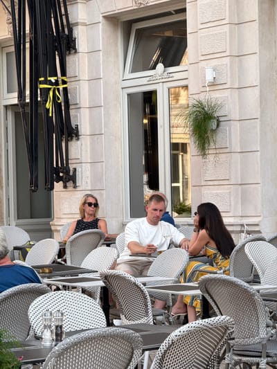 The image shows an outdoor seating area of a cafe or restaurant located on a street in Split, Croatia. In the foreground, there are several tables and chairs, characteristic of outdoor dining. The chairs appear to be made of wicker or a similar material with a grey and white woven pattern. On one of the tables, a salt and pepper grinder set is visible.
In the middle ground, four people are seated at tables. On the left, a blonde woman with sunglasses is sitting at a table. Next to her, a man in a white t-shirt is holding a glass of white wine. To the right of the man, a woman with long dark hair, wearing a yellow and blue striped dress, is seated. Further back and partially visible is another person. The building in the background is made of light-colored stone with large windows and ornate architectural details. A potted plant hangs from the building. In the upper left of the image, black metal umbrella stands are tied together with a yellow ribbon.