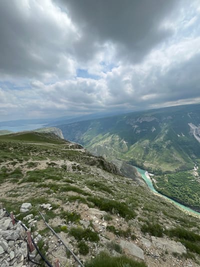 An elevated, angled landscape photograph focuses on a steep, grassy mountainside descending into a river valley. The sky is overcast with heavy, gray clouds, allowing glimpses of blue in patches. In the distance, a wide river flows through the valley, bordered by dense green forests and terraced slopes. The mountainside in the foreground is covered with sparse grass and rocky terrain, with exposed cables and metal supports near the bottom of the frame. The location is near the city of Dubki, Russia.