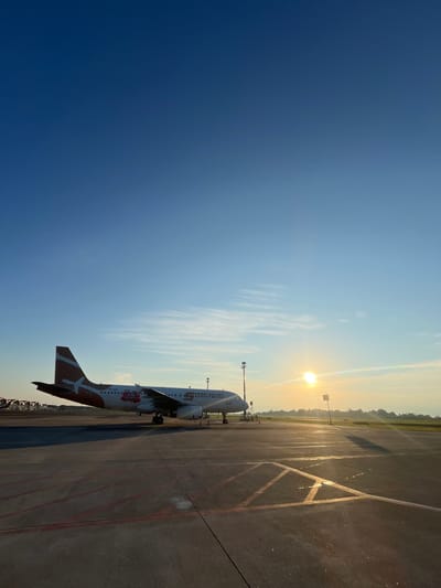 The image depicts a Super Air Jet airplane parked on an airport tarmac in Baji Mangngai, Indonesia. The sun is rising, casting a bright glare in the sky and shadows on the ground. The sky is clear blue, transitioning to a lighter shade near the horizon. The airplane is predominantly white with orange and red accents, including the tail. The tarmac shows markings and a strip of greenery runs along the edge of the airport.