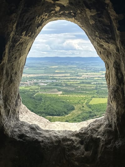 The image shows a view through a rock opening, possibly a cave or hollow. The rock is rough-textured and varies in color from light gray to dark shadow. Outside the opening, the landscape is a mix of green fields and forests, with patches of buildings and roads suggesting human settlement. The view is slightly rotated, with the horizontal plane appearing tilted. A strip of sky with clouds is visible above the landscape. Given the location provided, this could be a natural opening in rocks near the city of Han Krum, Bulgaria, providing a framed view of the surrounding landscape. No people or subjects are identifiable.