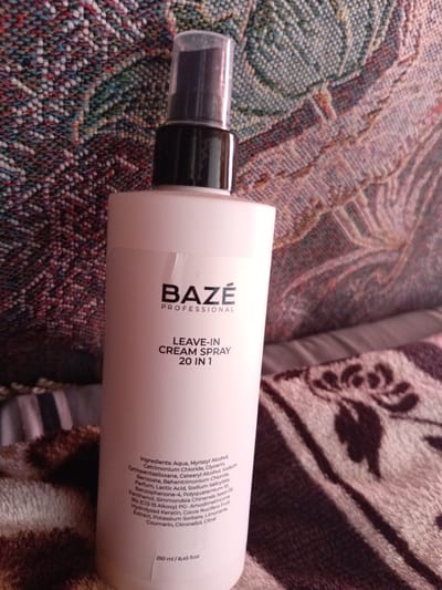 In Monastirische, Russia, a white bottle of "BAZE PROFESSIONAL LEAVE-IN CREAM SPRAY 20 IN 1" is placed on a patterned fabric surface. The fabric has a woven texture with a colorful, mottled design featuring shades of red, blue, green, and white. The bottle has a black pump top with a clear cap. Text on the bottle includes the brand name "BAZE PROFESSIONAL," the product name "LEAVE-IN CREAM SPRAY 20 IN 1," a list of ingredients, and the volume "250 ml / 8,45 fl.oz." The bottle is positioned slightly off-center to the right of the frame. The background is primarily the patterned fabric.