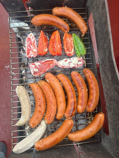 The image displays an overhead view of a charcoal grill in the city of Bangor, United Kingdom, actively cooking various food items. The grill grate supports multiple subjects. In the top section, three brown, cooked sausages are visible. Below these, five halved bell peppers are arranged: four red peppers and one green pepper, each filled with a white creamy substance, consistent with cheese or a similar dairy product. Further down, two irregular pieces of red meat, one topped with a white creamy substance, are situated next to two additional red bell pepper halves, also filled with the white substance. The lower portion of the grill grate holds ten sausages; two are lighter in color, appearing less cooked or a different variety, while the remaining eight are brown and cooked, displaying varying degrees of charring. Charcoal embers are visible beneath the grate, with aluminum foil present in some areas. The grill's exterior appears dark, with some red elements. To the left and bottom-left of the grill, a reddish-brown textured surface is visible, alongside a partial view of a dark shoe or boot in the extreme bottom-left corner. A dark gray vertical surface frames the right side of the image. No individuals are fully visible.